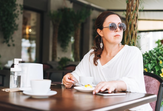 Brunette femme d'âge moyen à lunettes de soleil en vêtements blancs avec café à la terrasse du café d'été