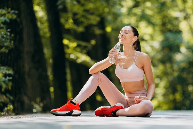 Brunette femme boit de l'eau dans les écouteurs assis dans le parc en plein air