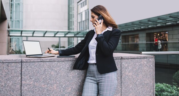 Brunette femme cadre debout et utilisant un ordinateur portable tout en parlant au téléphone à l'extérieur d'un bâtiment moderne