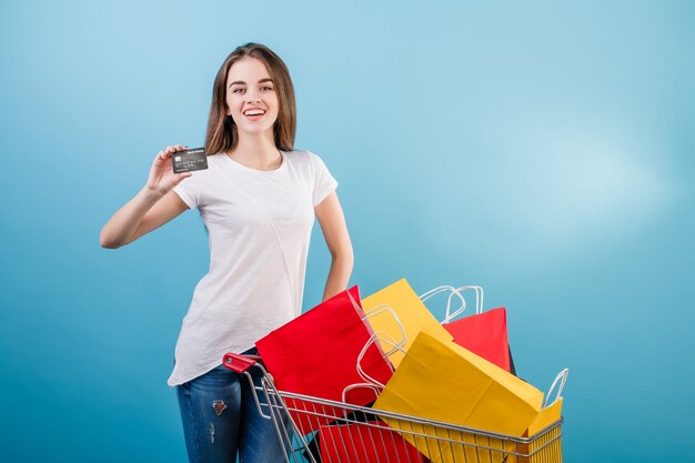 Brunette femme avec panier rempli de sacs en papier coloré et carte de crédit isolé sur bleu