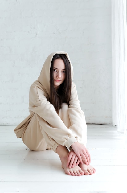 Photo brunette fille européenne en pantalons de survêtement et un sweat à capuche est assis sur un plancher en bois blanc dans une pièce lumineuse