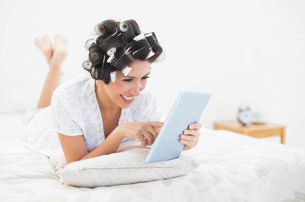 Brunette gaie dans les rouleaux de cheveux allongé sur son lit avec sa tablette