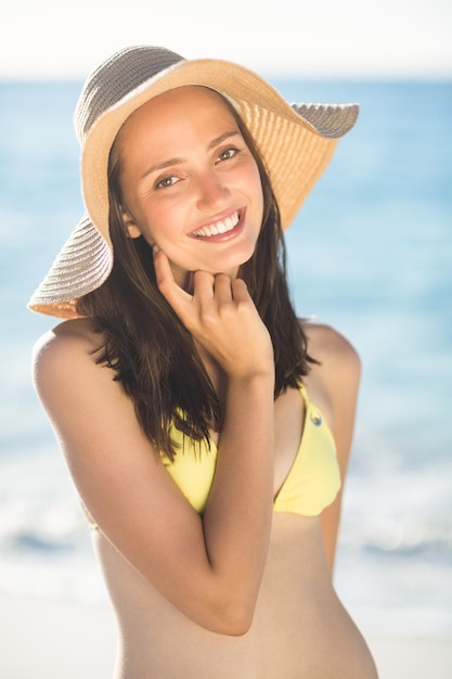 Brunette relaxante avec un chapeau de paille souriant à la caméra à la plage