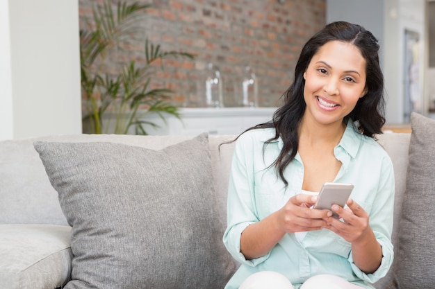 Brunette souriante à l&#39;aide d&#39;un smartphone sur le canapé