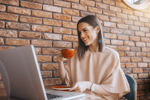 Brunette souriante habillée élégante de boire du café dans la cafétéria et regardant un ordinateur portable