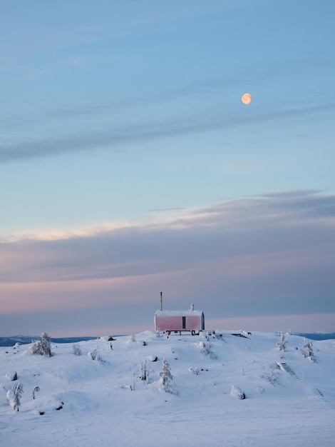 Bungalow solitaire et pleine lune sur une pente de montagne enneigée sous un ciel d'aube lumineux Fond minimaliste vertical avec une cabine rouge solitaire en hiver