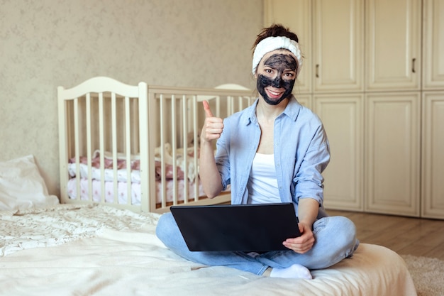 Bureau à domicile et fille indépendante dans des vêtements de maison d'un masque à charbon sur son visage travaille assis à la maison sur le lit.