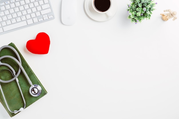 Bureau médical avec stéthoscope, livre, coeur, ordinateur portable, souris et tasse de café sur un bureau blanc.