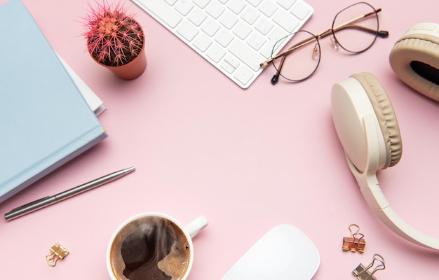 Un bureau rose pastel avec des écouteurs à café et des fournitures de bureau encadrant un espace vide