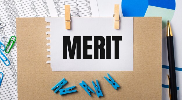 Sur le bureau se trouvent des rapports, des pinces à linge bleues et des graphiques, un stylo, un cahier et une feuille de papier avec le texte MERIT. Concept d'entreprise