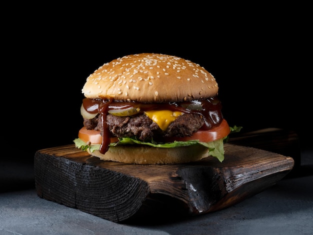 Burger avec bacon viande tomate et laitue sur planche de bois et fond noir gros plan