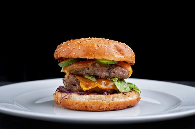 Burger avec double escalope de viande, fromage. concombre et laitue, sur une plaque blanche, sur fond noir