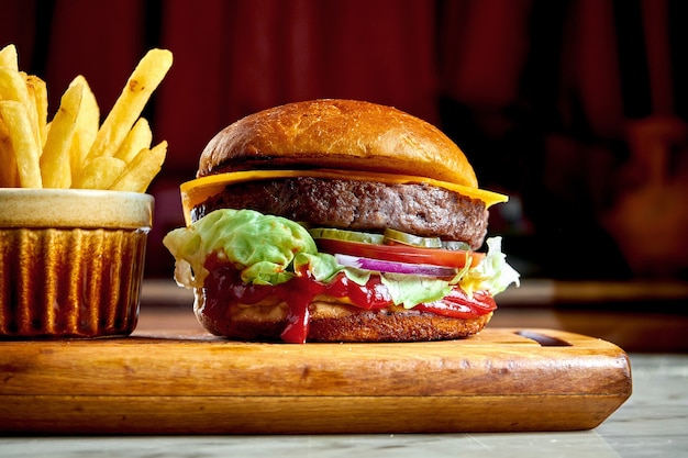 Burger juteux et appétissant avec tomates, laitue, concombre, fromage fondu et oignons avec pain brioché servi sur une planche de bois avec frites