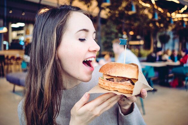 Burger en papier kraft dans les mains et la jeune fille mange avec ses mains dans un restaurant.