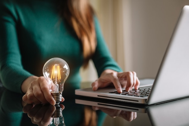 Business hand holding lightbulb with using laptop computer and money stack in office. idée d'économie d'énergie et de comptabilité concept de finance dans la lumière du matin