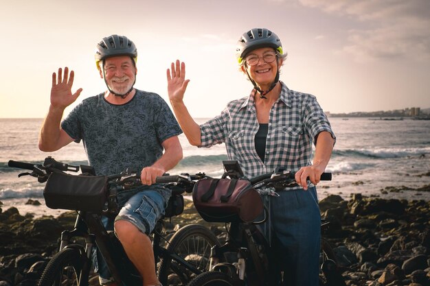 Bye bye Senior couple joyeux à cheval sur la plage de galets avec des vélos électriques au coucher du soleil Authentique vie à la retraite des personnes âgées et concept de mobilité durable