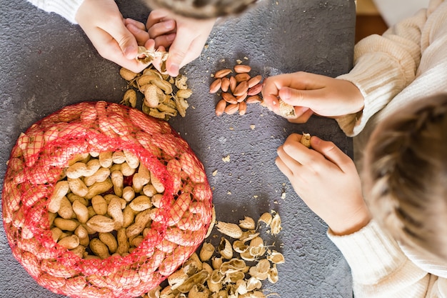 Cacahuètes, un sac en filet avec des noix non pelées et des cosses sur la table. Les mains des enfants pèlent des noix. Mode de vie. Vue de dessus