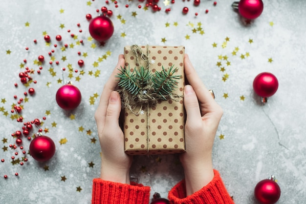 Cadeau d'artisanat en papier brun avec des brindilles de sapin de Noël et de la ficelle entre les mains d'une jeune fille vêtue d'un pull rouge.