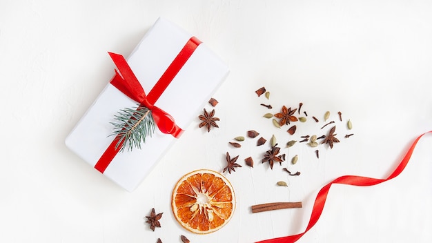 Un cadeau blanc attaché avec un ruban rouge se trouve sur un fond blanc entouré d'un ruban rouge, des tranches d'orange séchées, des bâtons de cannelle, de l'anis étoilé et des clous de girofle séchés. Flou artistique. Vue de dessus. Mise à plat. Noël