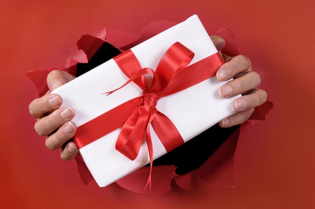Cadeau blanc avec ruban étant livré à travers un arrière-plan de papier déchiré rouge.