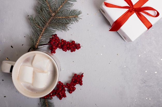 Cadeau blanc avec ruban rouge sur fond gris entouré de tasses à café avec mashmello et branches à côté de baies rouges. Concept de Noël. Mise au point douce