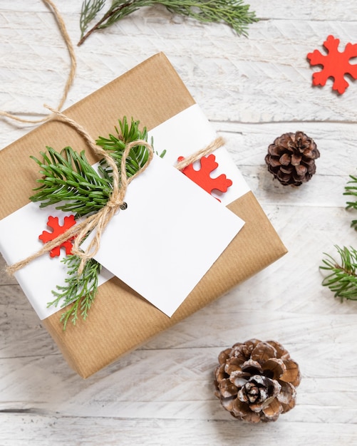 Cadeau enveloppé de Noël avec une étiquette-cadeau en papier carré sur une table blanche avec des branches de sapin et des décorations vue de dessus. Composition d'hiver rustique avec maquette d'étiquette-cadeau vierge, espace de copie