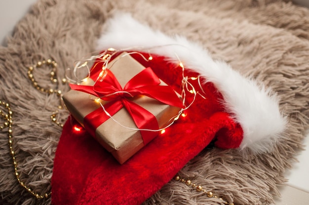 Photo un cadeau en papier kraft et avec un ruban rouge se trouve sur un chapeau rouge de noël sur fond de guirlande dorée