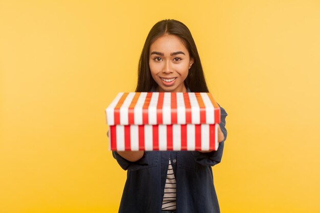 Cadeau pour toi. Portrait d'une généreuse fille brune heureuse avec un sourire charmant donnant une boîte à l'appareil photo, partageant un cadeau de vacances, félicitant pour son anniversaire. studio d'intérieur tourné isolé sur fond jaune