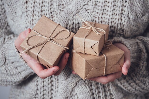 Cadeaux dans les mains des femmes, gros plan. Concept de cadeau monochrome et minimaliste. Une fille en pull tient des coffrets cadeaux en papier Kraft, attachés avec une ficelle. Fond de surprise, carte de voeux.