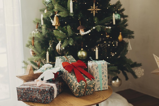 Cadeaux de Noël élégants dans du papier d'emballage festif avec des arcs et des ornements vintage sur une table en bois sur fond de lumières d'arbre de Noël doréesJoyeuses fêtes Réveillon de Noël atmosphérique