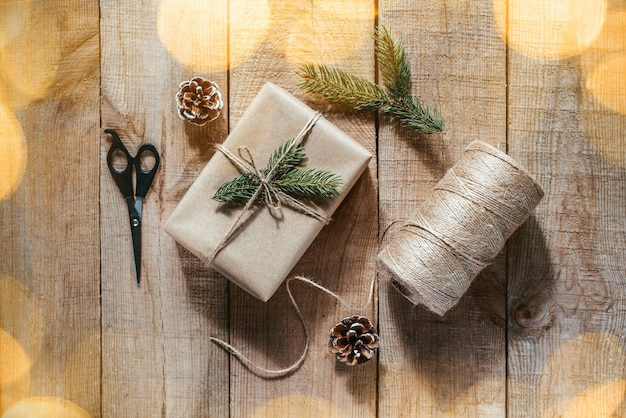 Cadeaux de Noël emballant des suppiles : boîte-cadeau, corde de jute, ciseaux, branches de sapin et pommes de pin sur fond de bois