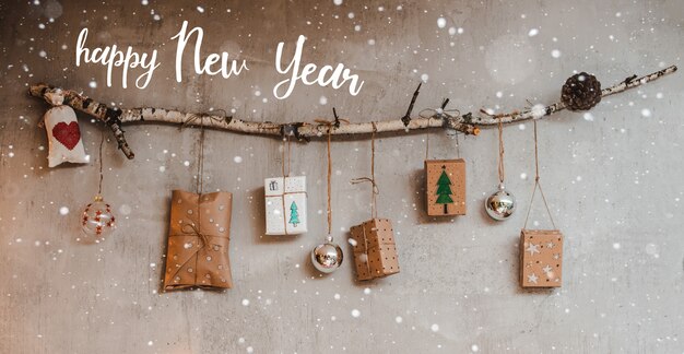 Cadeaux de Noël Emballés avec du papier Kraft et des décorations faites à la main, accrochés à une corde attachée à un bâton contre un mur de béton gris.