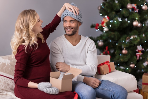 Cadeaux de noël. Joyeuse belle femme ravie assise sur le canapé et regardant son mari tout en l'aidant à mettre un chapeau chaud