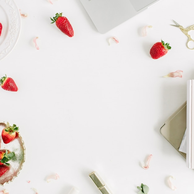 Cadre de bureau à domicile moderne et féminin avec ordinateur portable, ordinateur portable, rouge à lèvres, fraises crues fraîches et boutons de fleurs roses sur fond blanc. Mise à plat