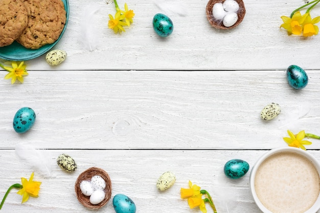 Cadre fait d'oeufs de Pâques, tasse de cappuccino, fleurs de printemps et biscuits sur table en bois blanc. composition de pâques