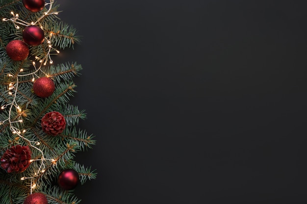 Cadre de Noël avec des branches de sapin, des boules rouges, une guirlande sur fond noir.