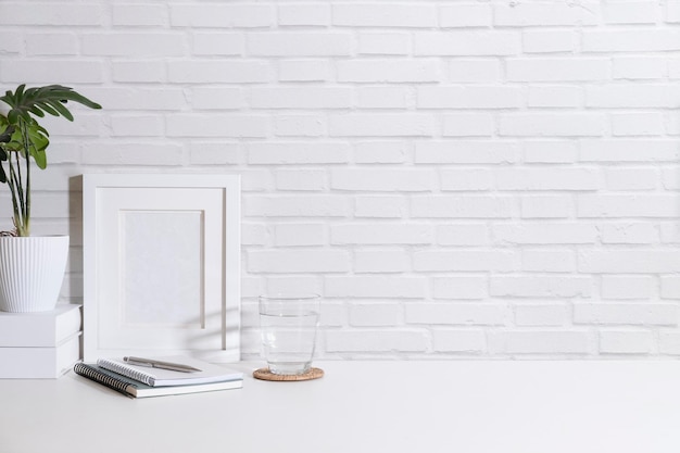 Cadre photo blanc plante en pot verre d'eau et ordinateur portable sur une table blanche contre un mur de briques Copiez l'espace pour votre texte publicitaire