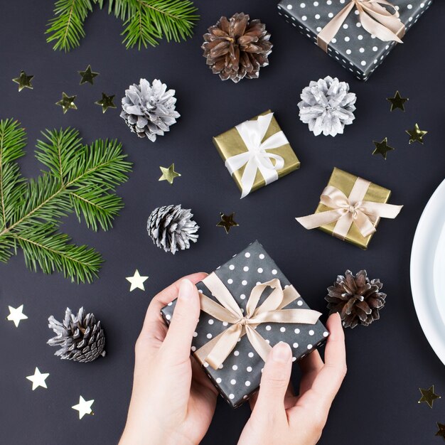 Cadre de table de Noël avec assiettes, cadeaux et décorations aux couleurs noir et or. Mains de femme tenant une boîte-cadeau. Vue de dessus