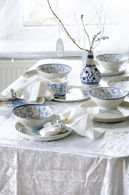 Cadre de table de Pâques élégant avec assiettes et bols de vaisselle en porcelaine peinte vide Décorations d'oeufs de Pâques branches dans un vase sur une nappe blanche devant la fenêtre Dîner de vacances de Pâques