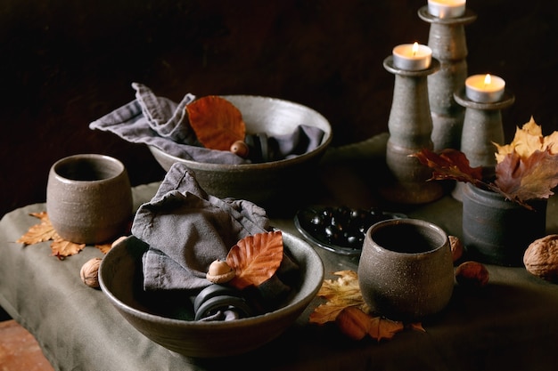 Cadre de table rustique d'Halloween ou de Thanksgiving d'automne avec de la vaisselle en céramique artisanale vide, des bols et des tasses gris, des bougies allumées sur une nappe en lin avec des feuilles et des glands jaunes d'automne. Fond sombre