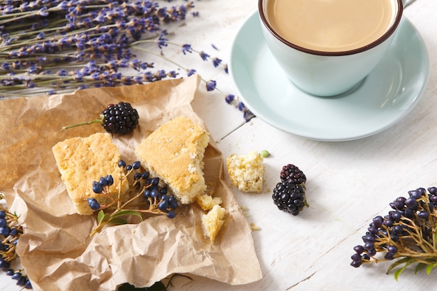 Café, biscuits, baies et composition de fleurs de lavande, gros plan sur bois blanc. Tasse bleue avec mousse crémeuse