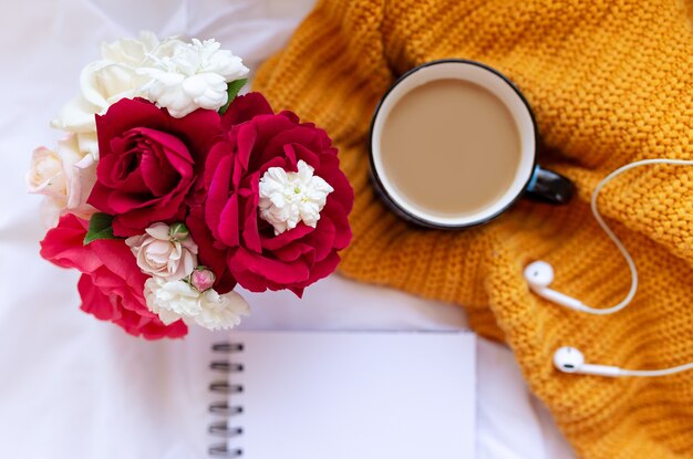 Café, cahier propre. fleurs roses, oreillettes sur des feuilles froissées blanches et vue de dessus de la couverture tricotée jaune. Femme travaillant à la maison. Petit déjeuner douillet. Maquette. Style plat.