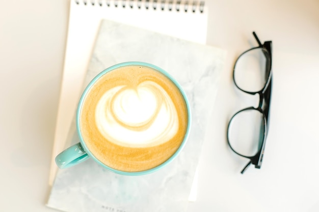 Café cappuccino dans une tasse bleue et un cahier de lunettes sur une table lumineuse au café Vue de dessus