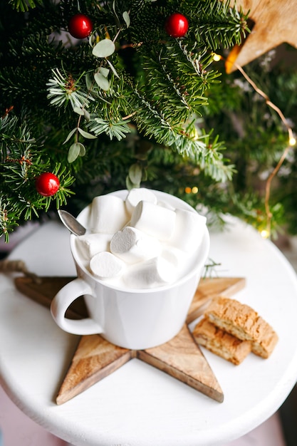 Café dans une tasse blanche avec des guimauves. Café de fête du matin avec des biscuits aux amandes cantuccini traditionnels italiens. Une tasse de café sur un fond de branches de sapin vert sur un support blanc.