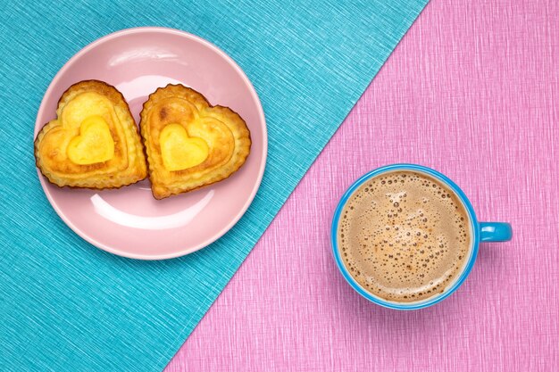 Café du matin et cupcakes en forme de coeur sur une nappe rose et bleue.