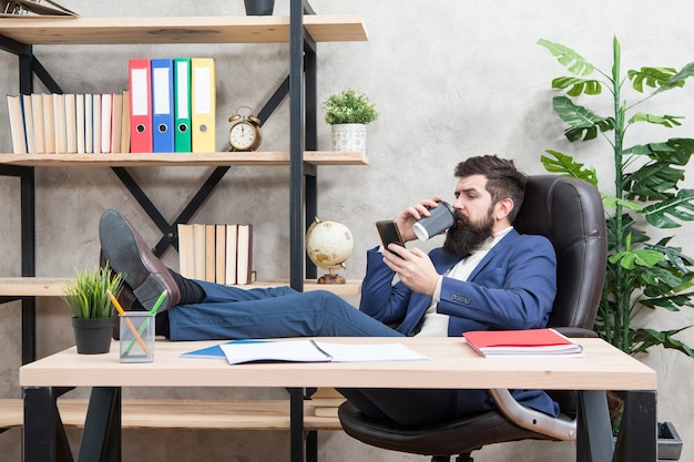 Le café est le gage de négociations réussies. Accro à la caféine. Appel mobile. Commencez la journée avec du café. Pause café détente. Patron profitant d'une boisson énergisante. L'homme d'affaires barbu tient la tasse et le smartphone.