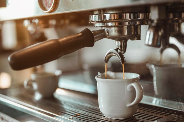 Café, extraction, profondeur, fabrication de machine, concept de barista. Cafetière en gros plan qui extrait professionnellement du café par un barista avec un verre blanc profond à la lumière du soleil du soir.