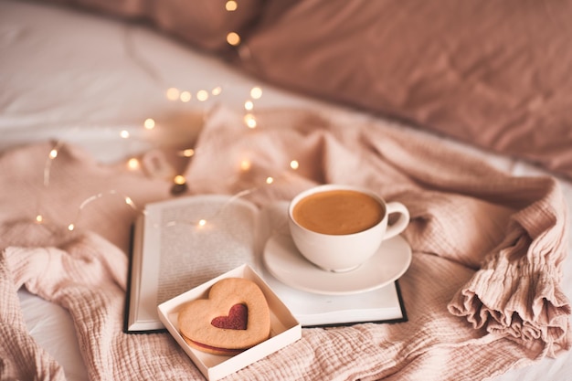 Café frais dans une tasse blanche restant sur un livre ouvert avec un gâteau en forme de coeur ou du pain d'épice sur un li brillant