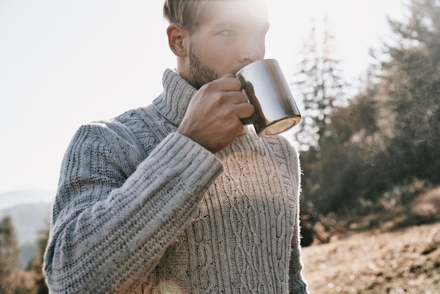 Le café frais a un goût délicieux. Beau jeune homme en pull chaud prenant le café du matin en camping dans les montagnes