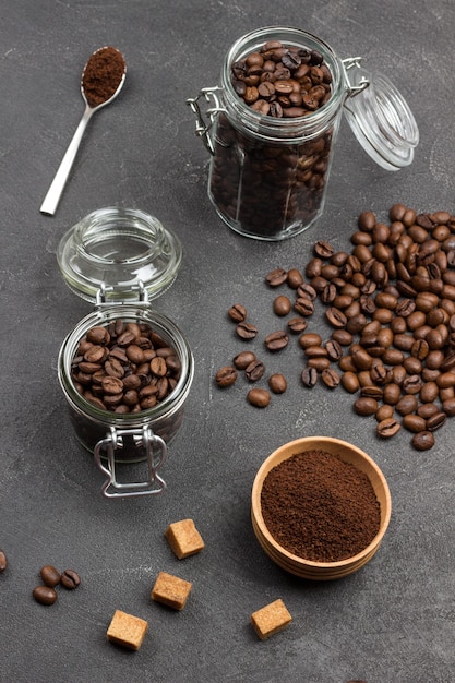 Café moulu dans un bol en bois et une cuillère. Grains de café torréfiés dans un bocal en verre et dans un sac en papier. Morceaux de cassonade sur table. Vue de dessus.